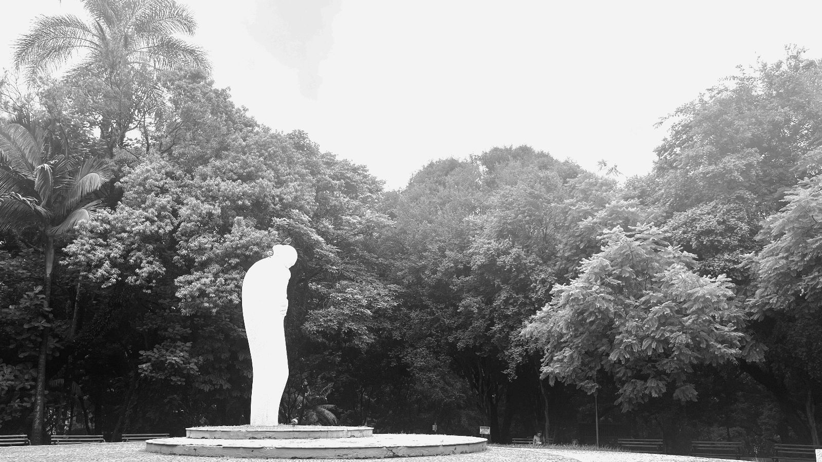 black and white photo of a clearing surrounded by dark trees. In the clearing, off center and elevated on 2 large flat discs is a marble figure, slightly hunched, crading something. The image has pretty high contrast, blowing out most of the details of the figure