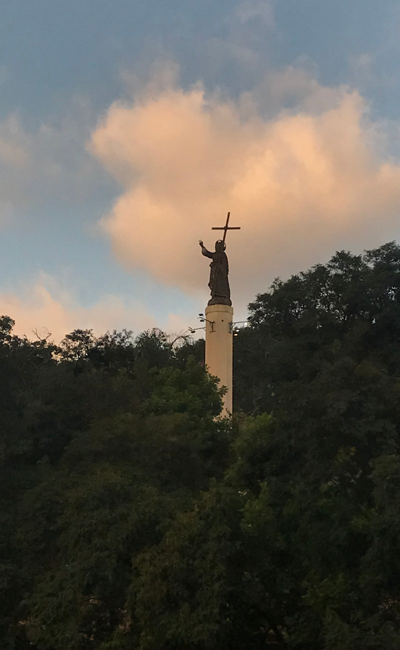 a statue of a saint stands above the treeline, the sky is approaching twilight, a golden cloud behind the figure