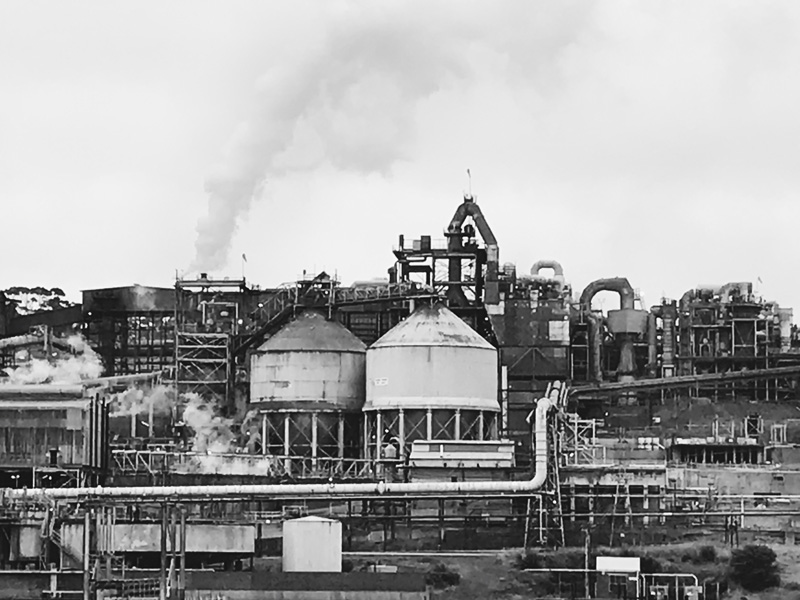 Black and white: A factory or processing plant with myriad pipes and some large tanks of unknown substance. There is a cloud of steam coming from a chimney