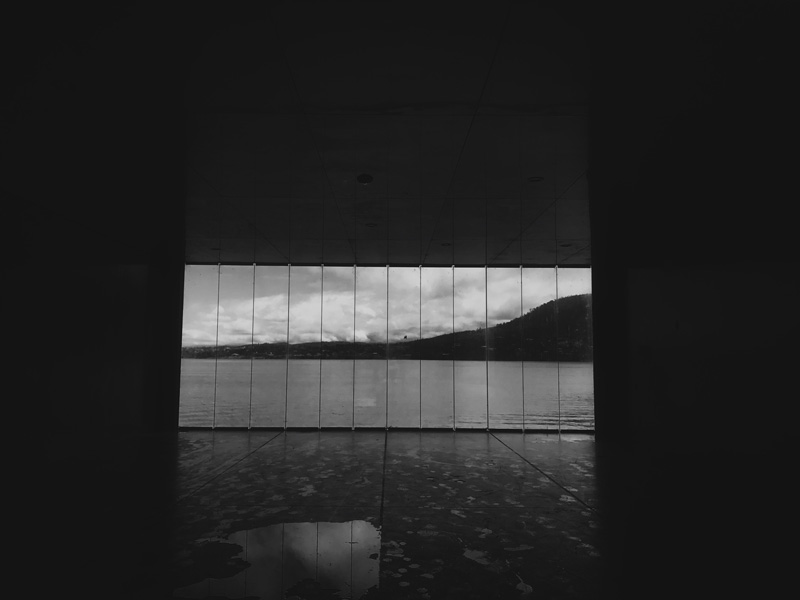 Black and white: A wide window looking across a harbour at a hill. There is a puddle in the foreground.