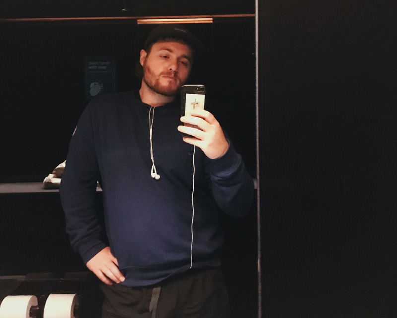 Colour: a selfie taken in low light at the mona bathrooms. I am wearing a blue sweater, and slightly pouting