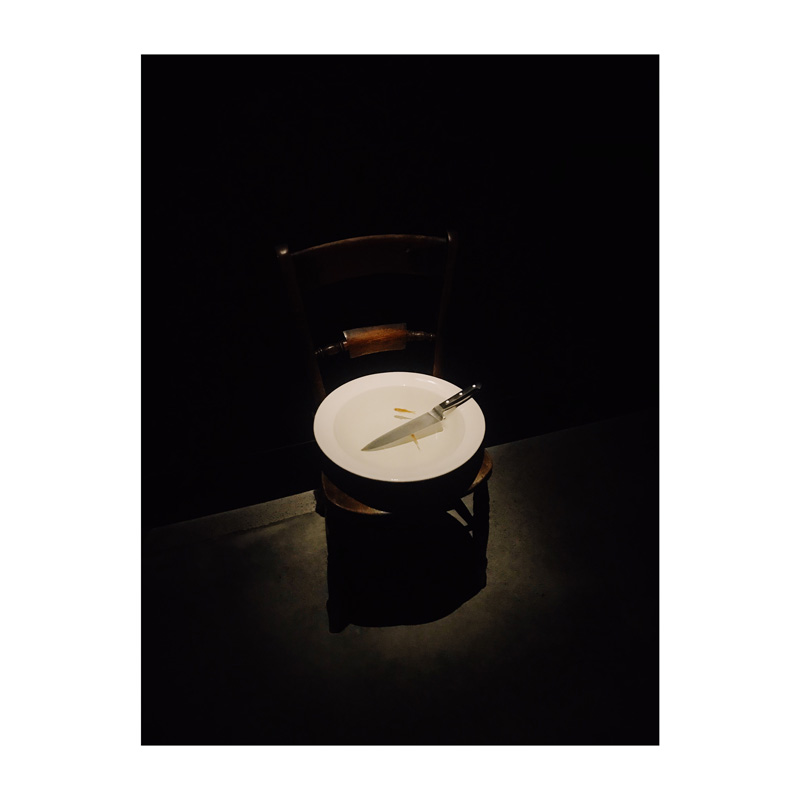 Colour: a very low lit photo of a bowl on a chair. Inside the bowl is water, a knife, and two goldfish